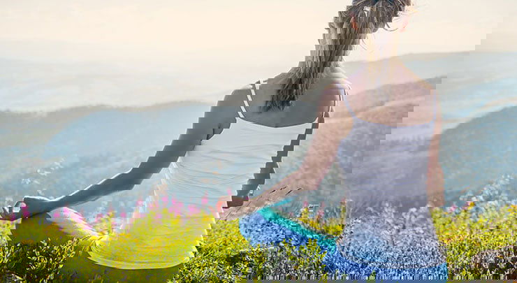 Woman in white tank top and blue pants sitting on grassy mountain meditating after clitoral hood reduction.
