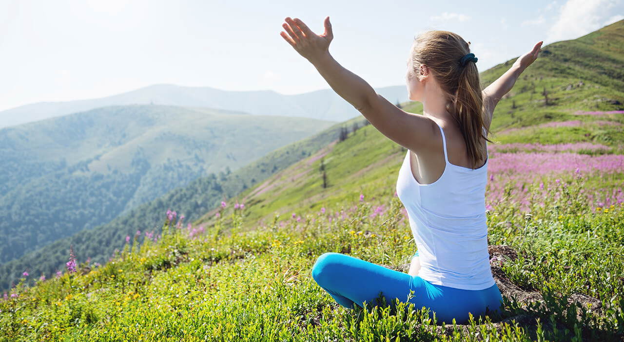 Woman sitting on mountaintop looking out with arms raised, inspired by her transformation.