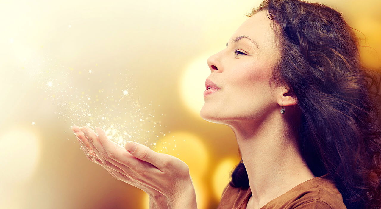Brunette woman holding up sparkles in her hands and blowing them while smiling.