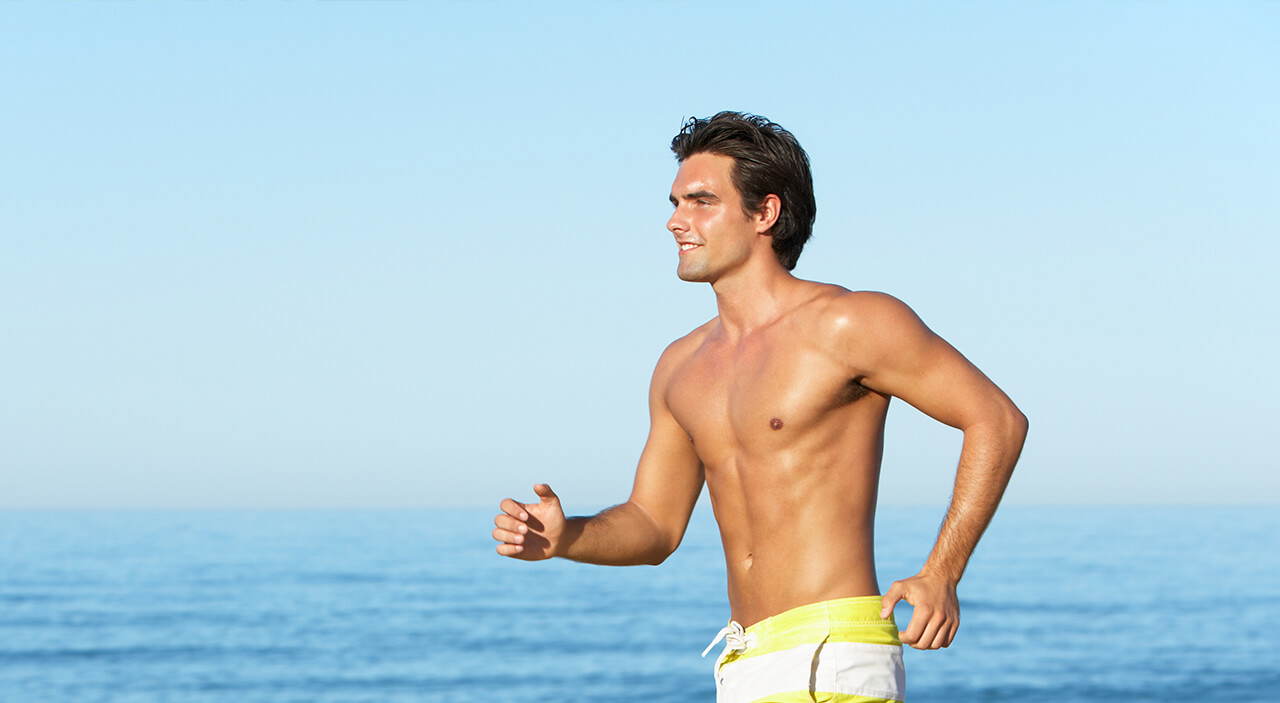 Confident man running shirtless on the beach after gynecomastia treatment.