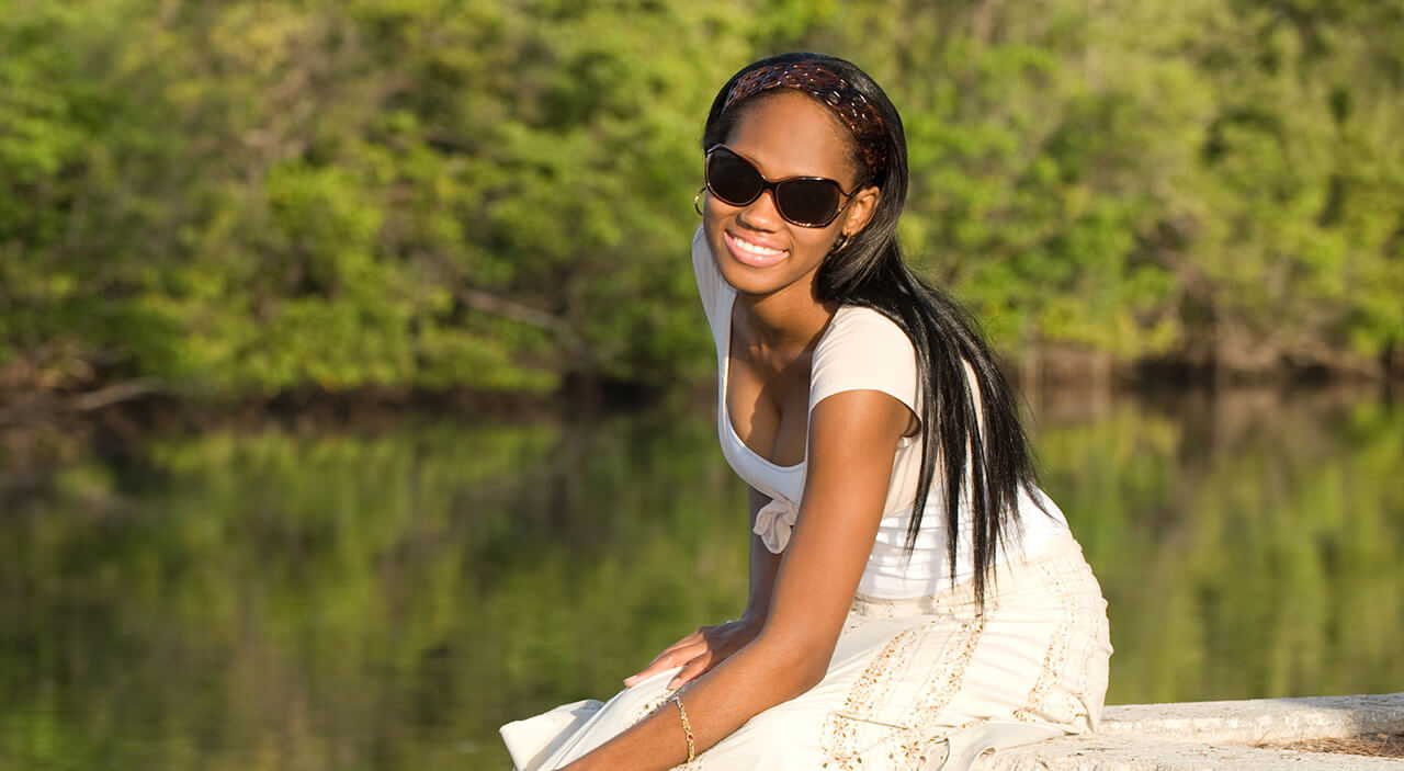 Woman sitting on dock at lake wearing white dress and sunglasses smiling.
