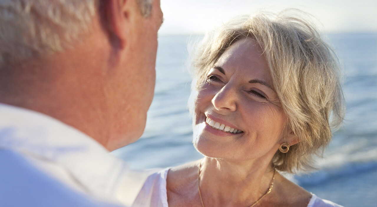 Radiant older woman smiling at her husband on the beach, looking naturally youthful.